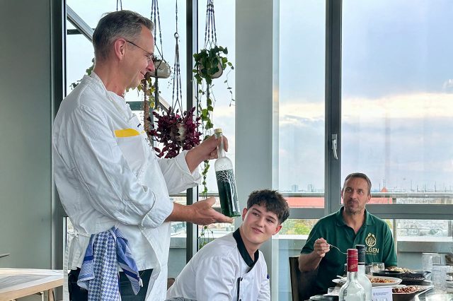 Saschs Kühnau berät an einem Tisch sitzende Personen. Er präsentiert dabei eine mit einem weißfarbenen Baum verzierte, Olivenölflasche aus Glas.