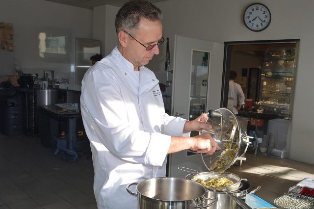 Sascha Kühnau in weißer Arbeitskleidung siebt gekochte Zutaten über einem Topf in einer professionellen Küche; im Hintergrund Edelstahlgeräte und ein weiterer Arbeitsraum.