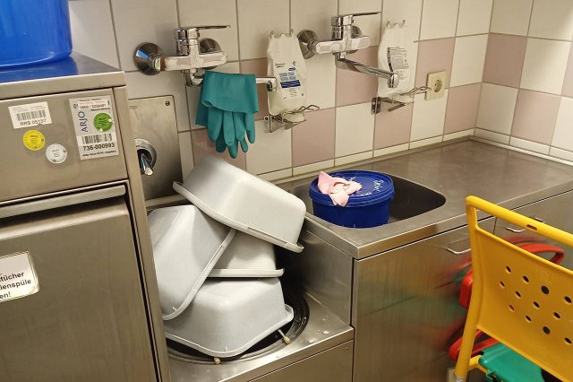 Spülbereich mit Edelstahlbecken, übereinandergestapelten Kunststofftabletts, Wandarmaturen, Einmalhandschuhen und einem Reinigungseimer in einer hauswirtschaftlichen Umgebung.