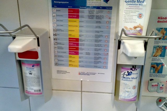 Wandmontierte Spender für Händedesinfektion und Waschlotion neben einem Reinigungs- und Hygieneplan an einer gefliesten Wand.