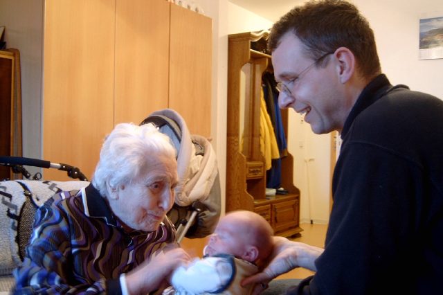Schascha Kühnau in dunklen Hemd unterstützt eine ältere Darme mit weißen Haar im Umgang mit einem Baby. Lockere, entspannte Stimmung in einem einfachen Wohnzimmer.