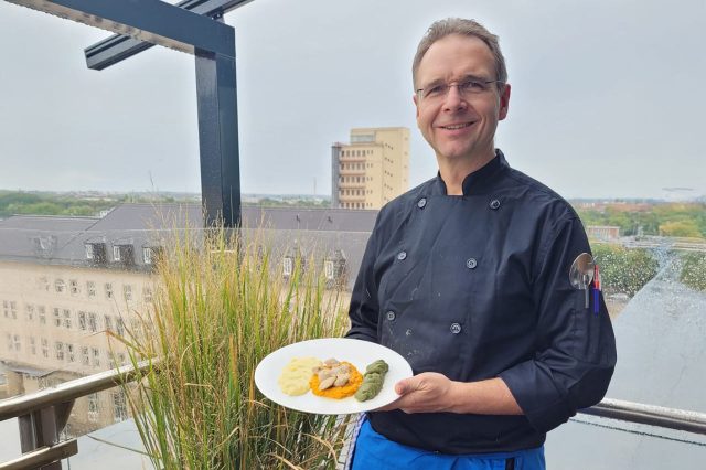 Sascha Kühnau in schwarzer Kochjacke steht im Freien auf einer Terrasse und hält einen Teller mit angerichtetem Gericht; im Hintergrund Stadtansicht und bepflanztes Geländer.