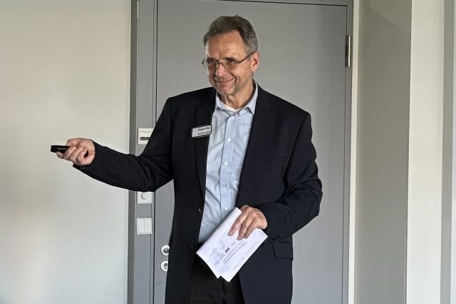 Sascha Kühnau im Anzug während eines Seminars. Er hält Unterlagen in der Hand und lauscht mit amüsierter Geste einem Teilnehmer.