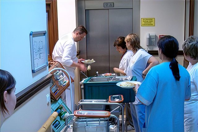 An einem Speisenwagen demonstriert Sascha Kühnau in Kochkleidung anwesenden Pflegefachkräften Handgriffe im Umgang mit Lebensmitteln. Sie stehen im Flur einer klinischen EInrichtung.