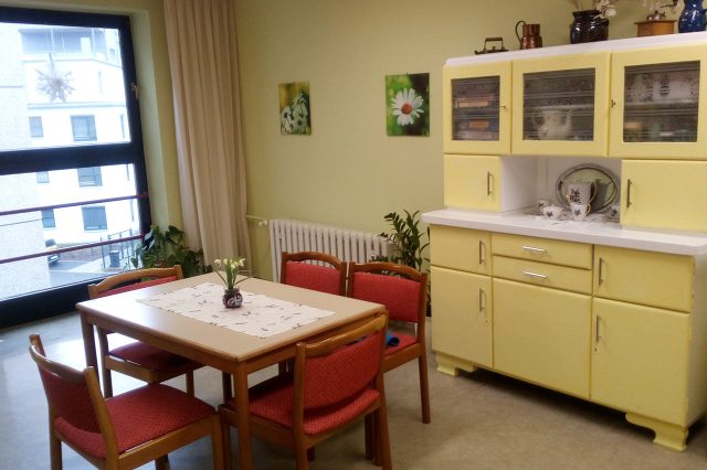 Heller Aufenthalts- oder Essraum mit Tisch und vier Stühlen, gelbem Buffetschrank, Wandbildern mit Blumenmotiven und großem Fenster mit Blick nach draußen.