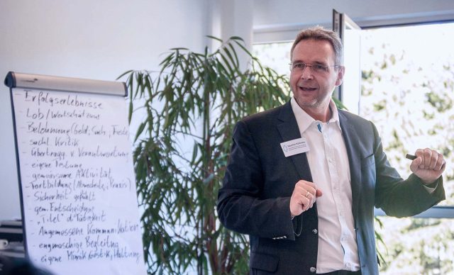 Sascha Kühnau in einem legeren Anzug gekleidet hält neben einem beschrifteten Whiteboard ein Beratungsgespräch mit engagierter Körpersprache.